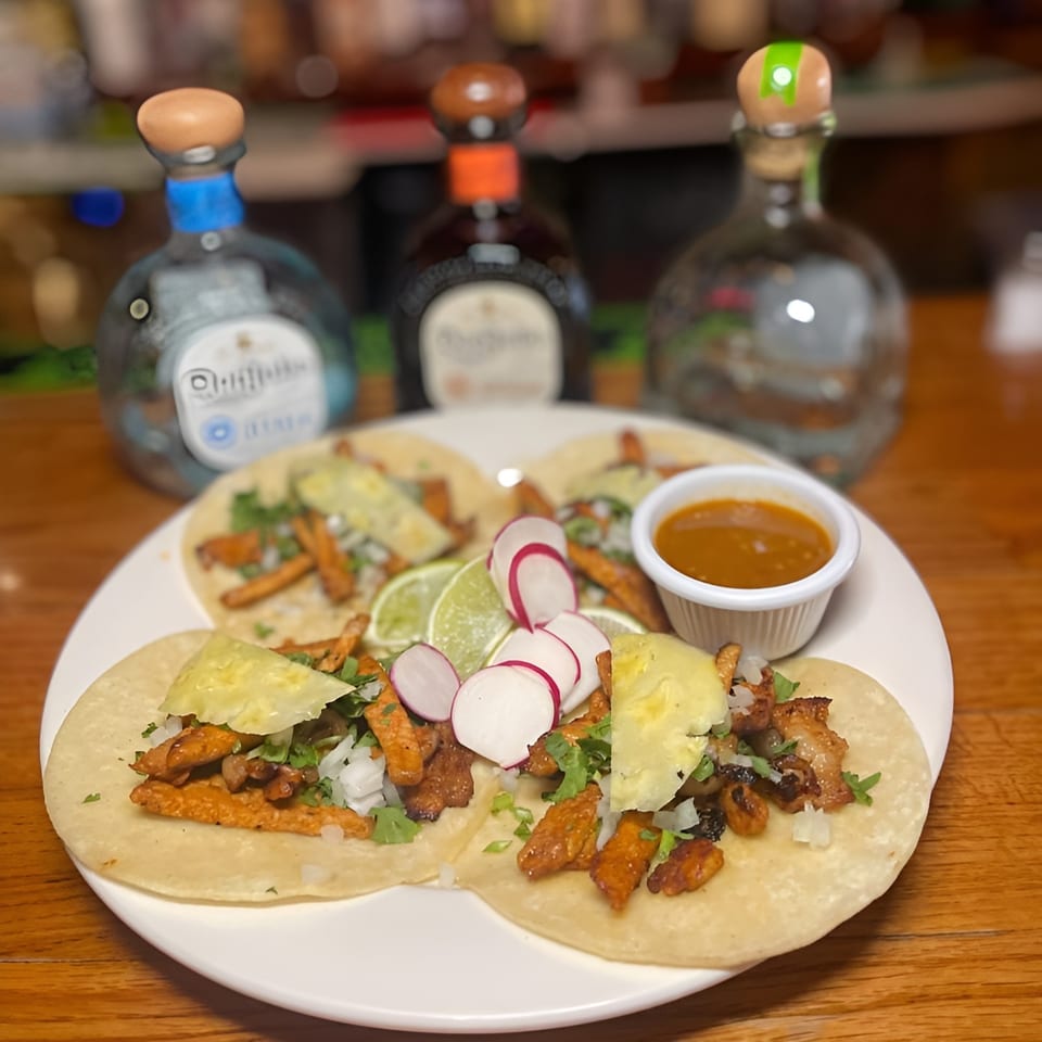Four Tequila's 55 tacos with pineapple, radish, and lime, served with sauce and tequila bottles.