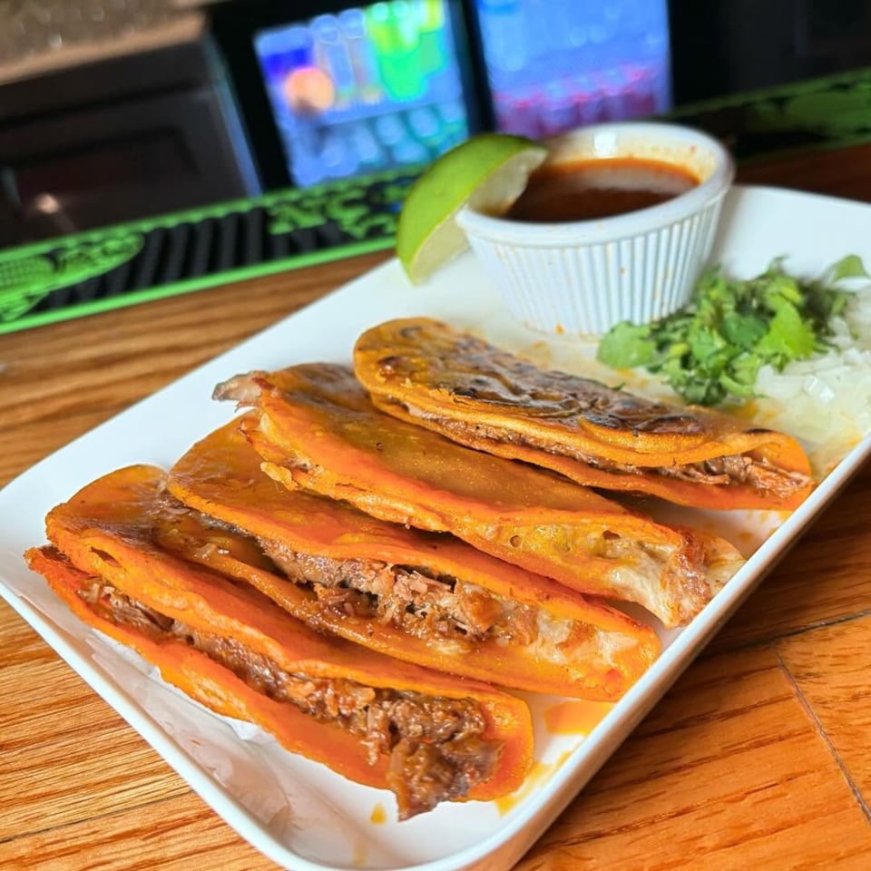Four birria tacos with a side of consommé, cilantro, and onions on a white plate.