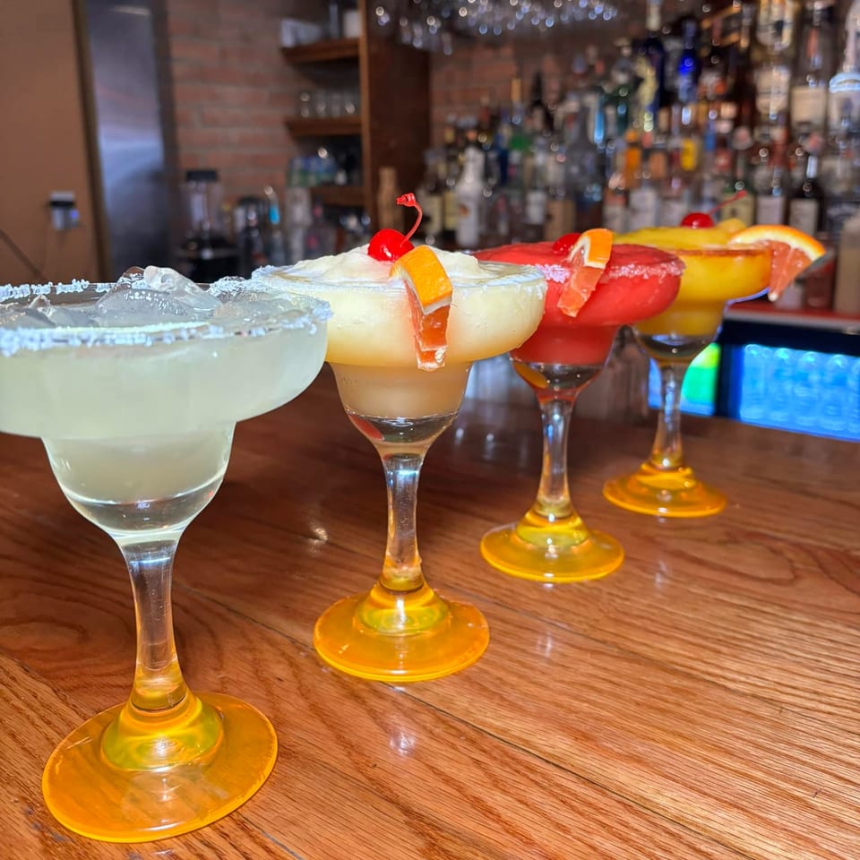 Four colorful margaritas with fruit garnishes and salt rims sit on a wooden bar at Tequila's 55.