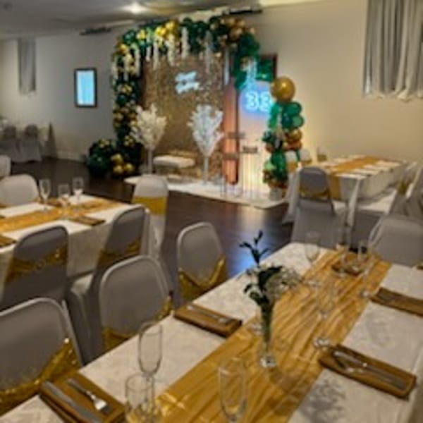 A banquet room with tables set with gold runners and a decorated stage with balloons and neon signs.