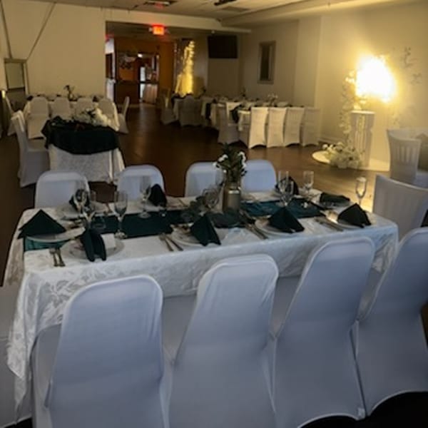 A long dining table set with white tablecloths, dark green napkins, and place settings in a banquet room.